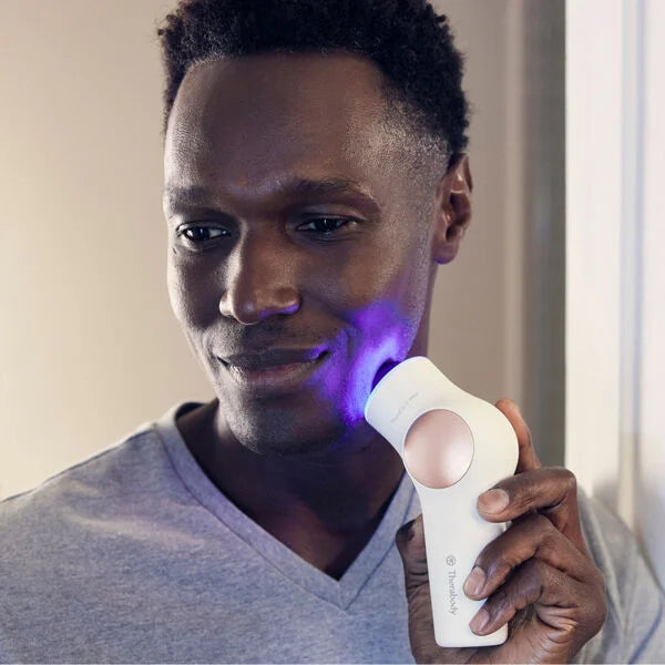 Black man with slight smile wearing gray t-shirt using TheraFace PRO facial massage device with blue light therapy on his face.